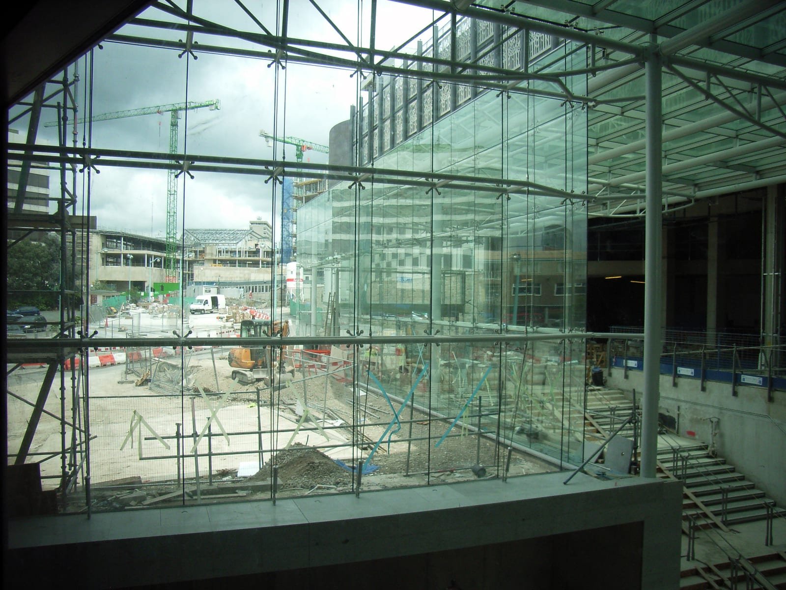 Illustrative image: a shopping centre under construction, seen through a glass wall, with cranes and earthworks outside.